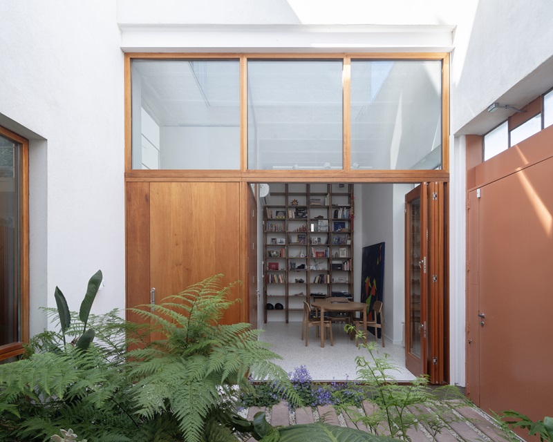Estudio-Damero-Casa-Planes: patio de entrada ajardinado con puerta de madera hacia el estudio