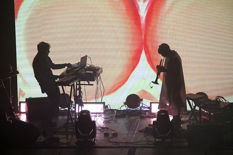 Imagen de dos músicos actuando en el escenario con visuales de color naranja.