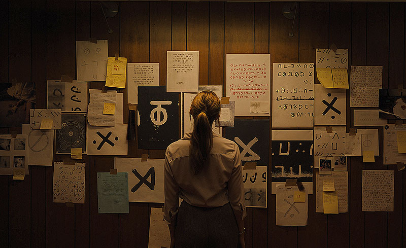 Longlegs - fotograma de la película, se ve a una chica mirando un panel en la pared lleo de símbolos raros