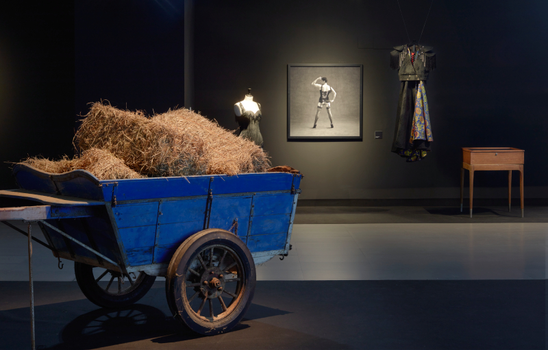 Vista de la exposición con un carro de paja e imágenes de fondo.