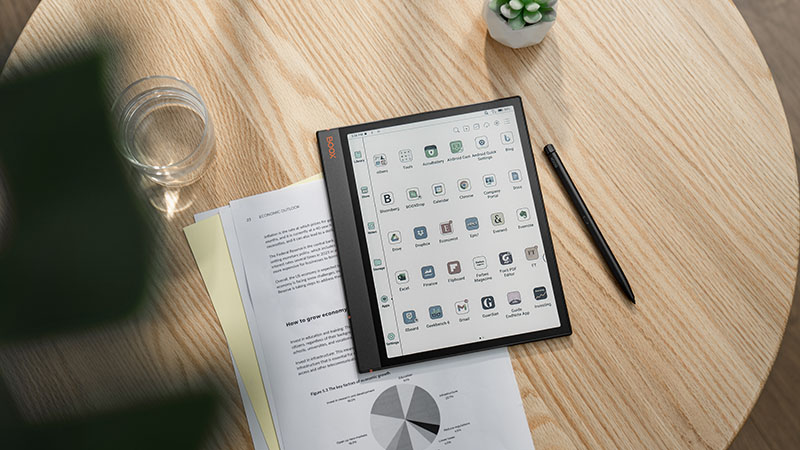 Boox Note Air3 C: la pantalla encendida muestra las distintas aplicaciones a color que tiene instaladas. Está sobre unos apuntes impresos en blanco y negro y a su vez sobre una mesa de madera que tiene un vaso de agua, una planta pequeña y el bolígrafo del dispositivo.