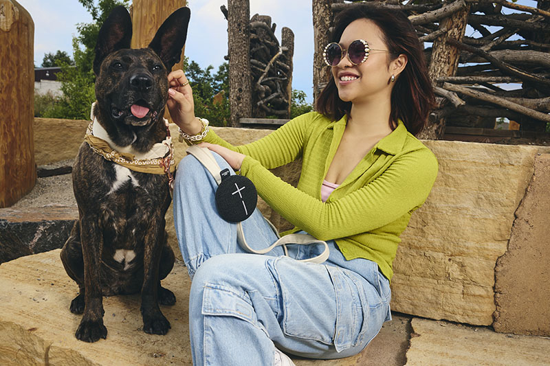 Miniroll: una mujer con gafas de sol y sentada en un banco de piedra con un perro oscuro mediano acaricia al animal, mientras escucha sonriente el sonido que emite el altavoz negro de Miniroll, que está enganchado a la correa de su bolso