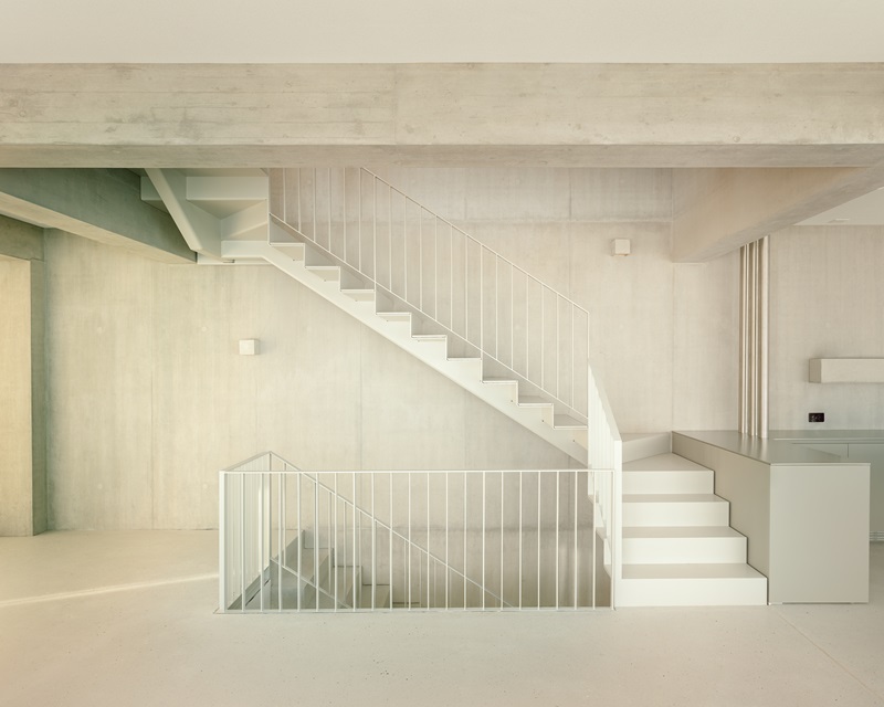 Oberwil-House-Caesar-Zumthor: sala de estar con vigas y pilares de hormigón y escalera metálica blanca