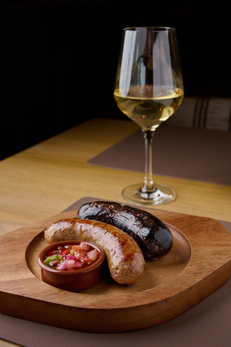 Al Fuego Madrid: salchicha parrillera y una morcilla argentina, dispuestas en una tabla de madera, junto a una copa de vino blanco