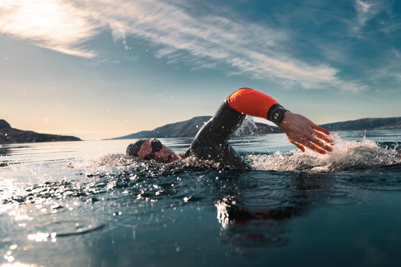 Fēnix 8: un hombre nada a crol en un pantano al atardecer. Lleva gafas y gorro de buceo y un reloj inteligente de Garmin que mide su actividad física.