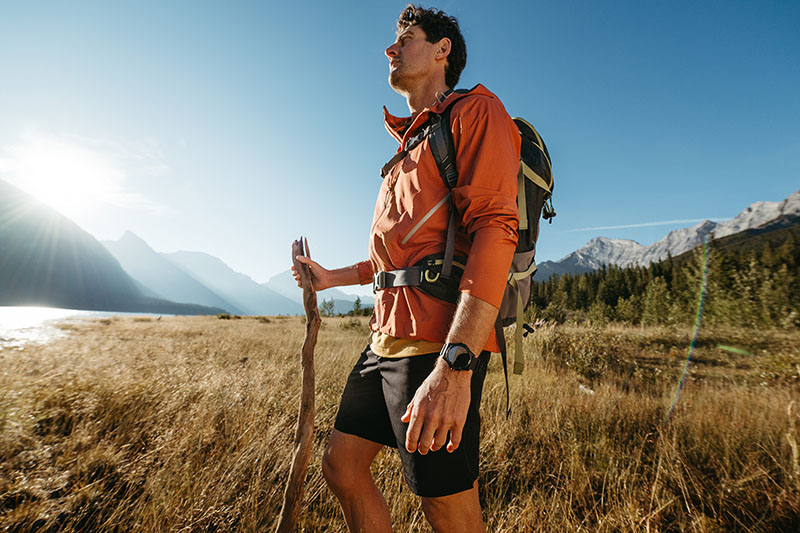 TicWatch Atlas: plano americano de un joven de piel clara y pelo moreno con un cortavientos naranja y una mochila de montaña haciendo senderismo con un palo grande por un paisaje espectacular un día soleado