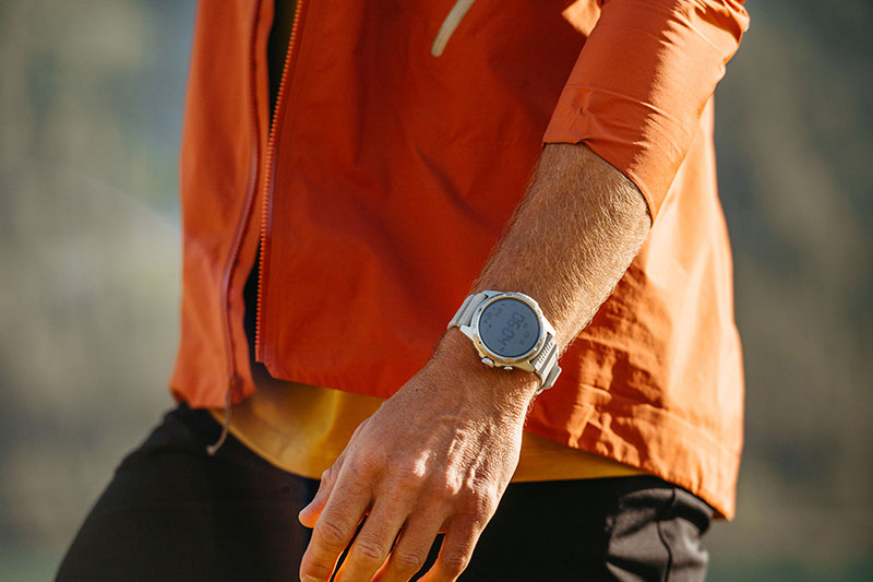 TicWatch Atlas: plano medio de un torso masculino con pantalón negro, cortavientos naranja y muñeca con un TicWatch Atlas plata al aire libre un día soleado