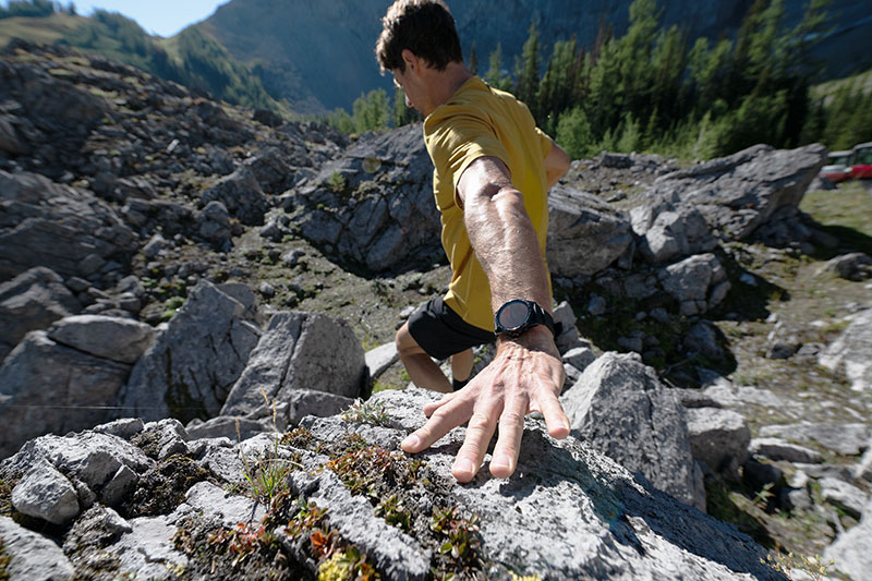 TicWatch Atlas: un hombre camina por la montaña un día soleado y se apoya en una roca al bajar con un TicWatch Atlas negro en su muñeca