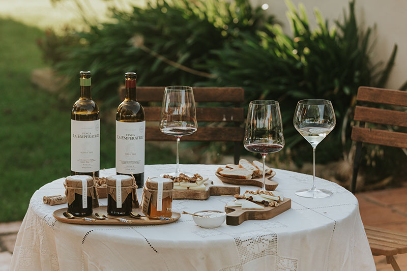 Finca La Emperatriz: Una mesa decorada con un mantel blanco, dos botellas de vino (tinto y blanco 2018) de Finca La Emperatriz, copas servidas y pequeños platos con mermeladas, nueces y otros aperitivos