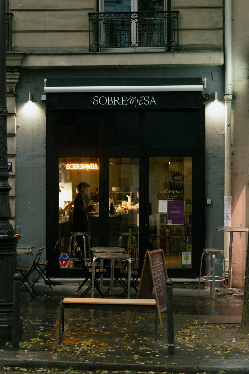 Entrada del restaurante Sobremesa en París, con un letrero iluminado que se asoma desde un ambiente lluvioso