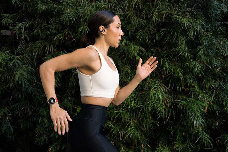 Amazfit Active: sobre un fondo de árbol natural se ve el plano medio de una mujer vestida con mallas negras y top blanco que va corriendo con su reloj Amazfit Active 2.