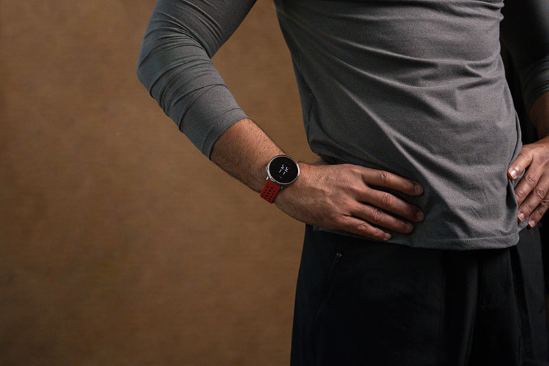 Amazfit Active: sobre un fondo de en tono tierra se ve el torso de un hombre con pantalón negro y camiseta gris deportiva en reposo con las manos en la cintura, la derecha con el reloj Amazfit Active 2 con correa roja de silicona.