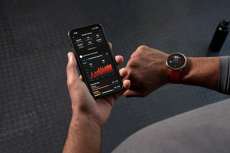 Amazfit Active: sobre un suelo negro de gimnasio se ven las manos de un hombre que sujeta un dispositivo móvil encendido con los datos que proporciona el reloj Amazfit Active 2 con correa roja de silicona que lleva en la otra.