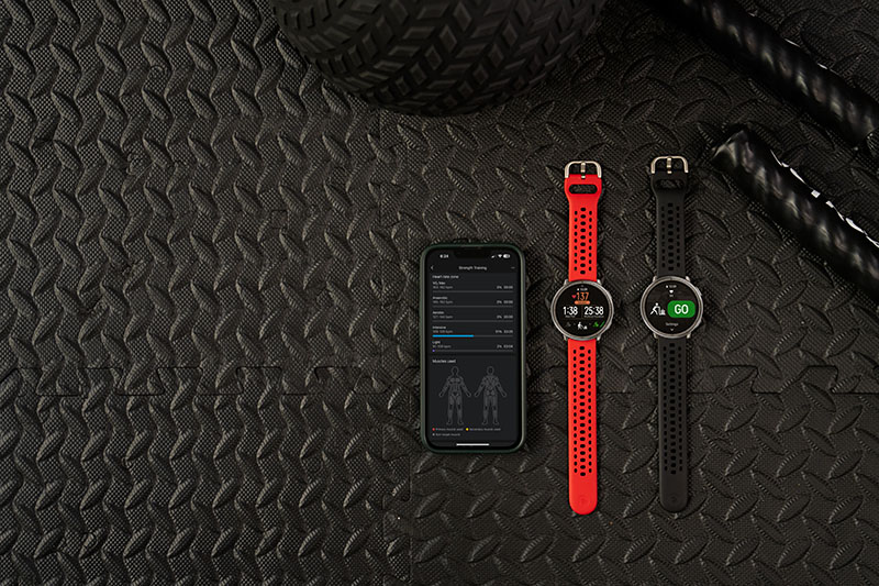 Amazfit Active: sobre un suelo negro con textura se ve un dispositivo móvil y dos modelos del reloj inteligente Amazfit Active 2, que son iguales, pero se distinguen tanto por los datos que muestran en la pantalla como por el color de la correa (roja y negra).