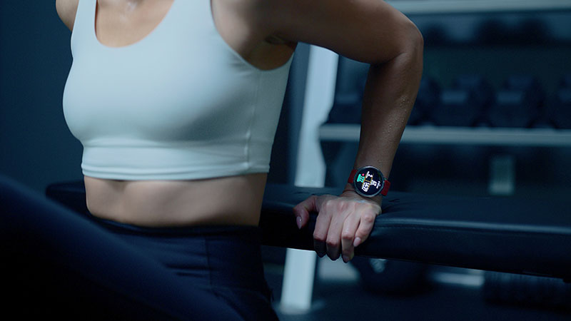 Amazfit Active: sobre un fondo de gimnasio difuminado se ve el torso de una mujer con mallas negras y top blanco realizando un ejercicio de tríceps con el reloj Amazfit Active 2 encendido.