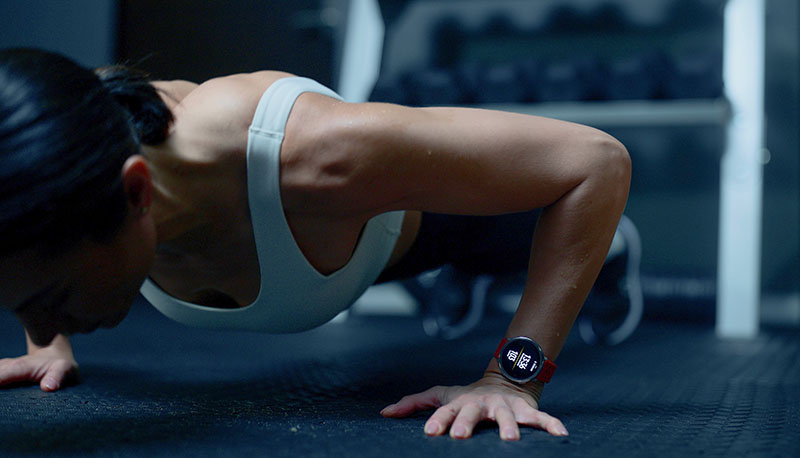 Amazfit Active: sobre un fondo de gimnasio difuminado se ve a una mujer vestida con mallas negras, top blanco y zapatillas de deporte haciendo flexiones con el reloj Amazfit Active 2 encendido.