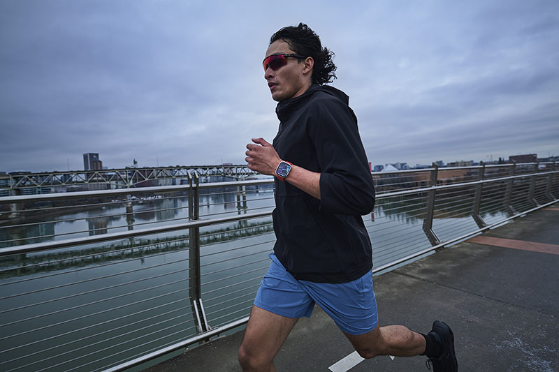 Amazfit Bip 6: un hombre entrena corriendo al aire libre con el reloj Amazfit Bip 6 en su mano izquierda. Lleva puesto calzado negro, pantalón corto azul, sudadera negra y gafas de espejo rojas.