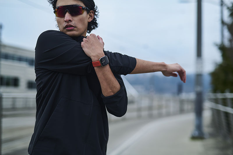 Amazfit Bip 6: un hombre realiza estiramientos al aire libre con el reloj Amazfit Bip 6 en su mano izquierda. Lleva sudadera negra y gafas de espejo rojas.