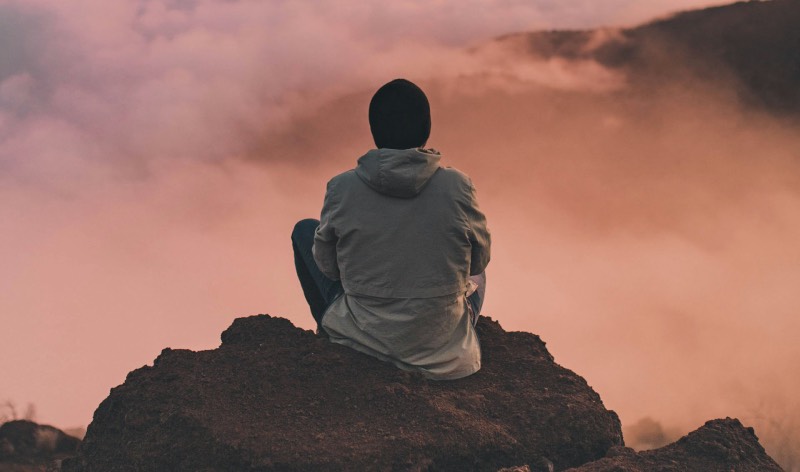 Opera Air: un chico sentado de espaldas sobre una roca de montaña contempla un atardecer de colores anaranjados entre las nubes.