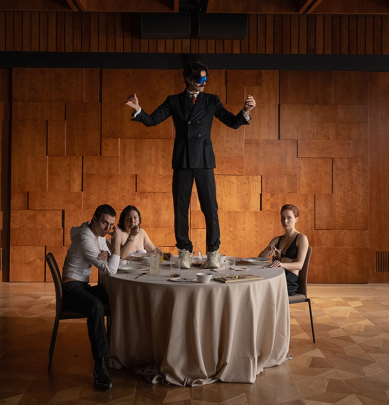 Ernesto Artillo. imagen de la performance "Tregua" se ve a unos personajes sentados al rededor de una mesa y a otro subido