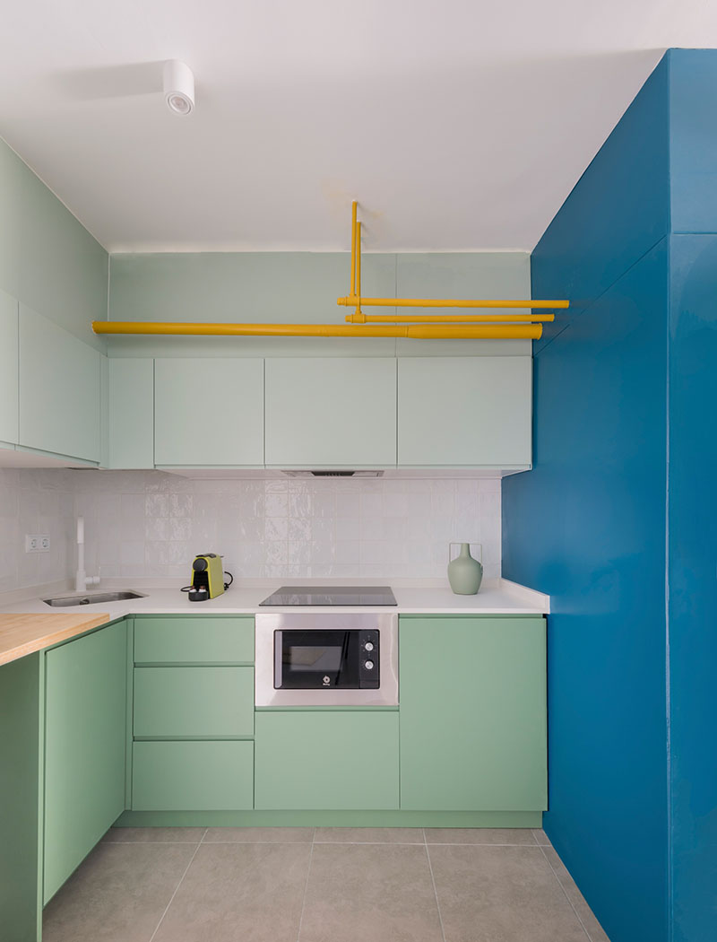 Ooiio Arquitectura: cocina compacta en tonos verdes y azul, con tuberías amarillas en el techo perteneciente a uno de los apartamentos en Carabanchel