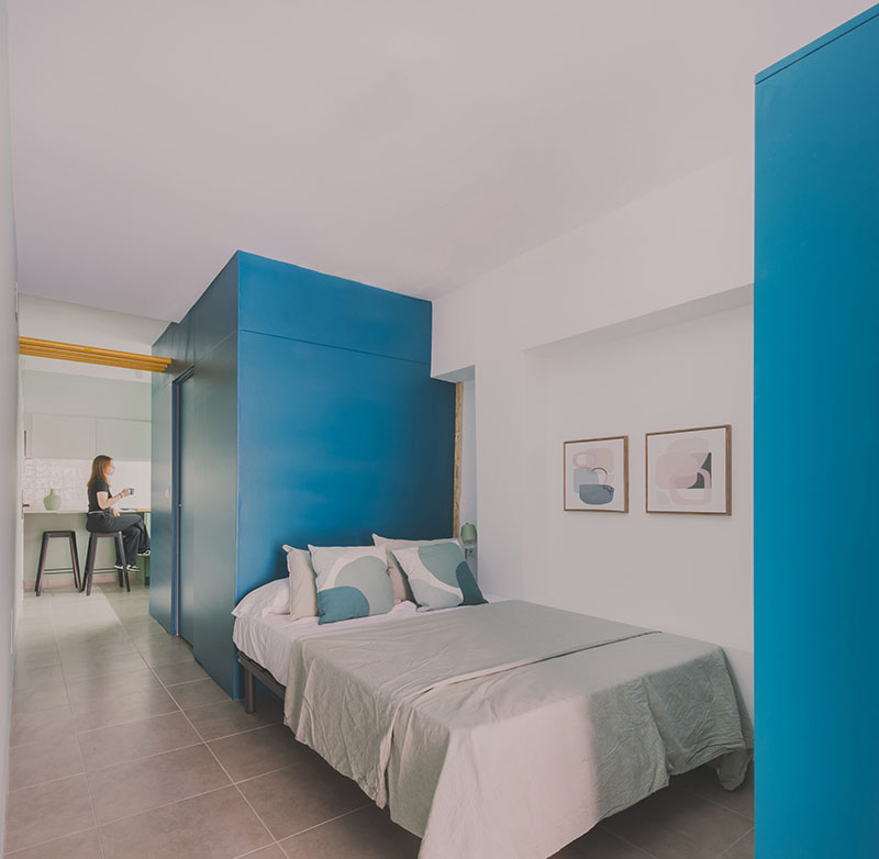 Ooiio Arquitectura: dormitorio con cabecero azul intenso y acceso directo a la zona de cocina perteneciente a uno de los apartamentos en Carabanchel